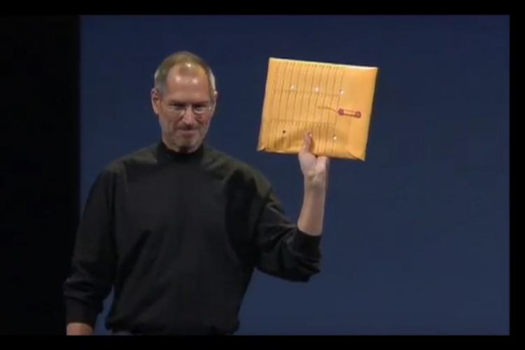 Steve Jobs vestido con su habitual jersey de cuello alto de color negro y sosteniendo con su mano izquierda un clásico sobre de color marrón donde se suelen guardar documentos de tamaño folio. 